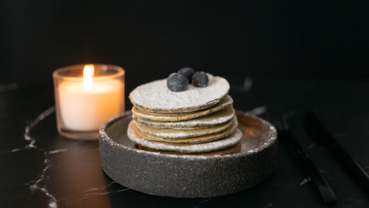 Stack of tasty pancakes with powdered sugar and blueberries served in bowl near burning candle on table against black background